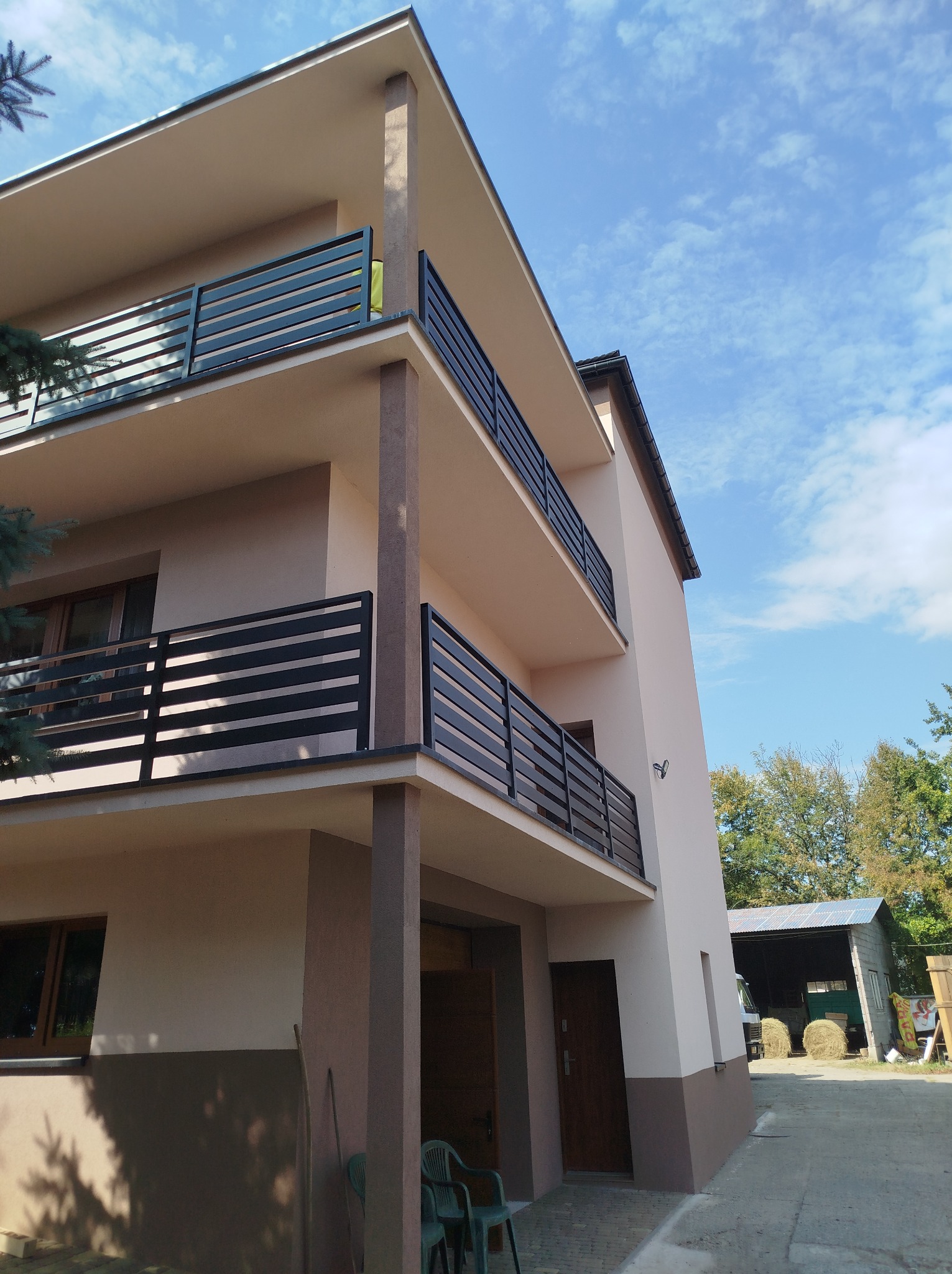 Trzykondygnacyjny budynek z balkonami zabezpieczonymi czarnymi, poziomymi balustradami, widok z dołu na tle błękitnego nieba z chmurami.