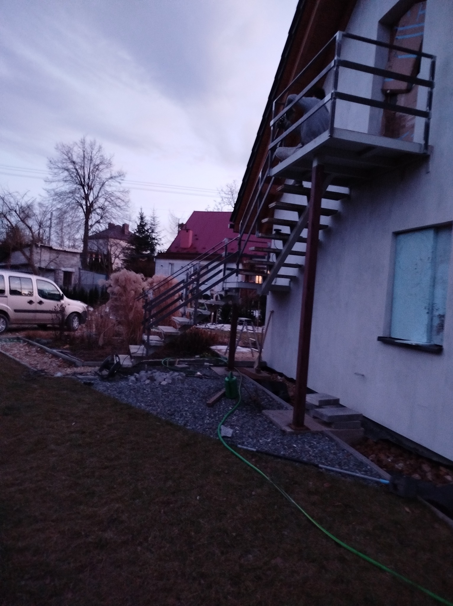 Zewnętrzne schody z metalową konstrukcją prowadzące na balkon domu jednorodzinnego, widok z ogrodu w pochmurny dzień.