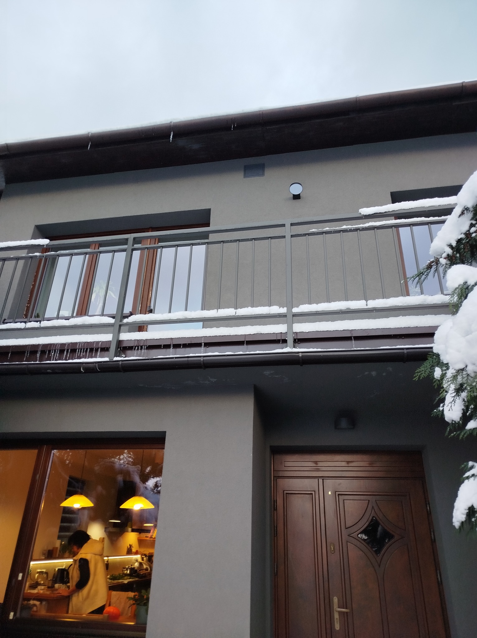 Szara elewacja domu z balkonem i ciemnobrązowymi drzwiami wejściowymi. Na balkonie metalowa barierka pokryta śniegiem, zwisające sople lodu. W oknie widoczna osoba w kuchni.