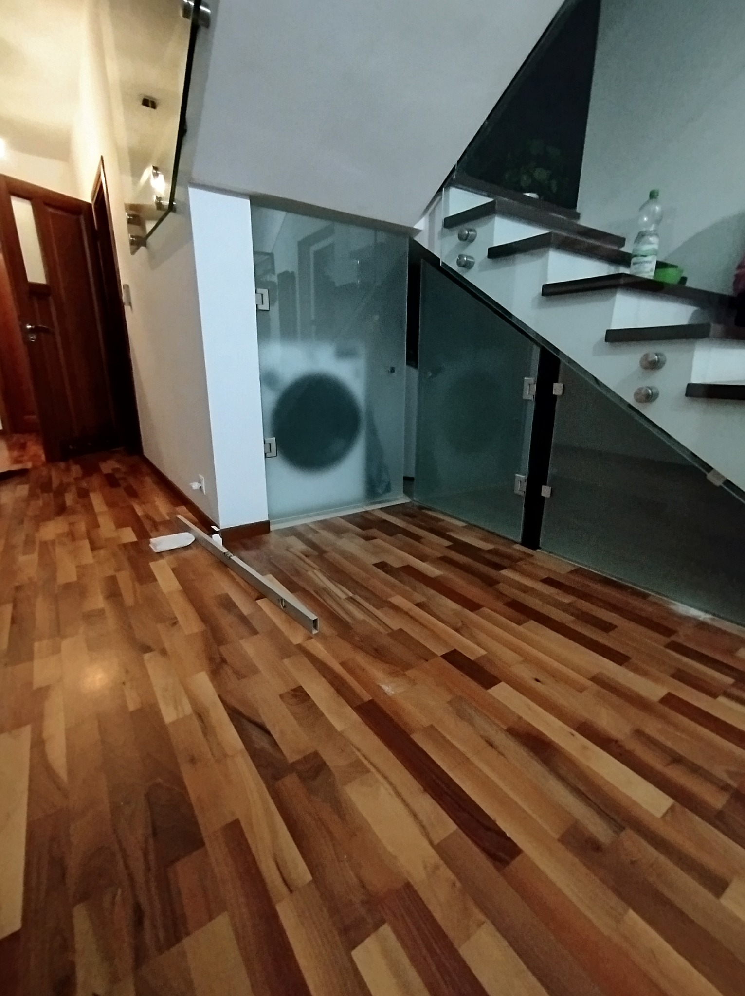 Szklana zabudowa pod schodami z widoczną pralką w tle, drewniana podłoga w trakcie montażu, poziomica leżąca na parkiecie.