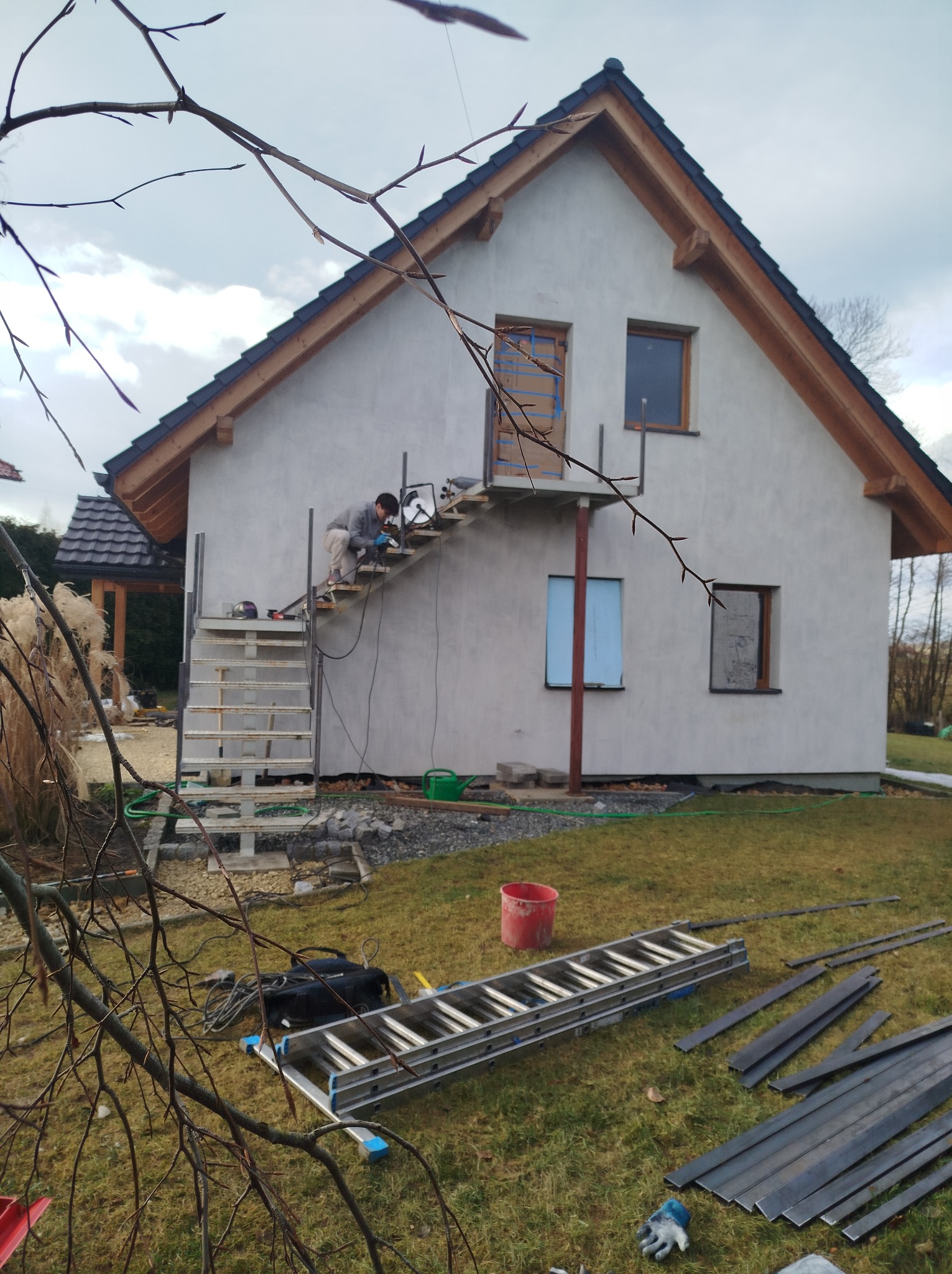 Pracownik w trakcie montażu metalowej konstrukcji schodów zewnętrznych przy ścianie budynku, widoczne narzędzia i materiały budowlane na trawie, w tle elementy ogrodu i fragment dachu