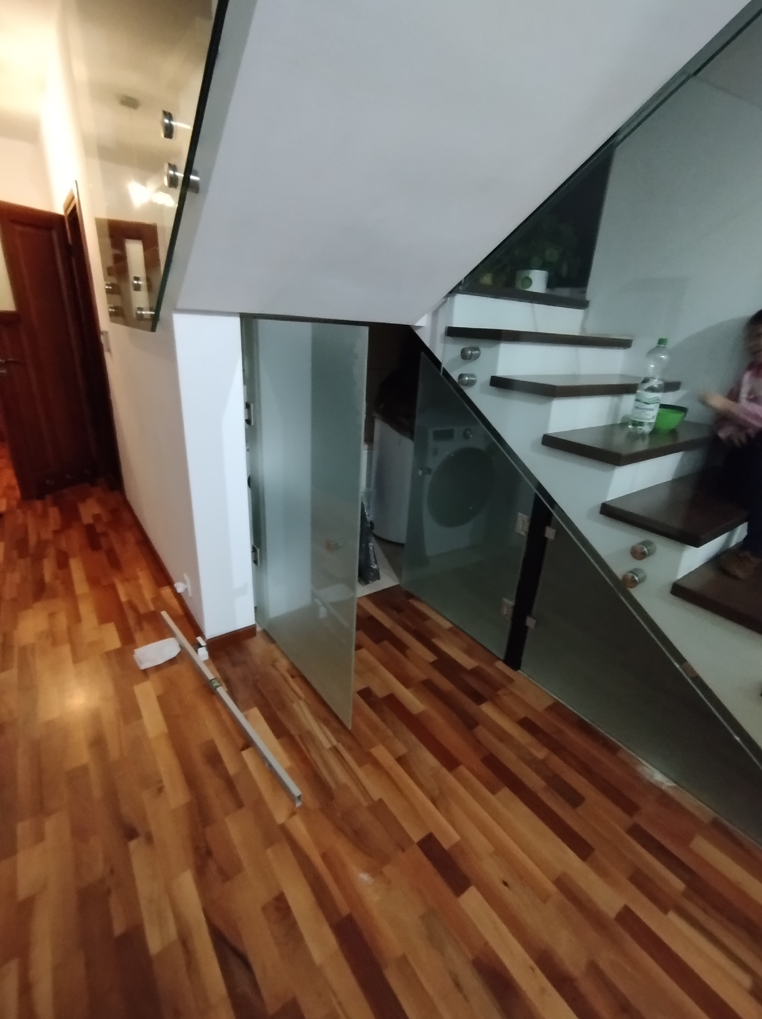 Szklana balustrada schodowa z widocznymi mocowaniami punktowymi, drewniane stopnie schodów, pralka w tle, drewniana podłoga, pomiarowe narzędzia na podłodze.