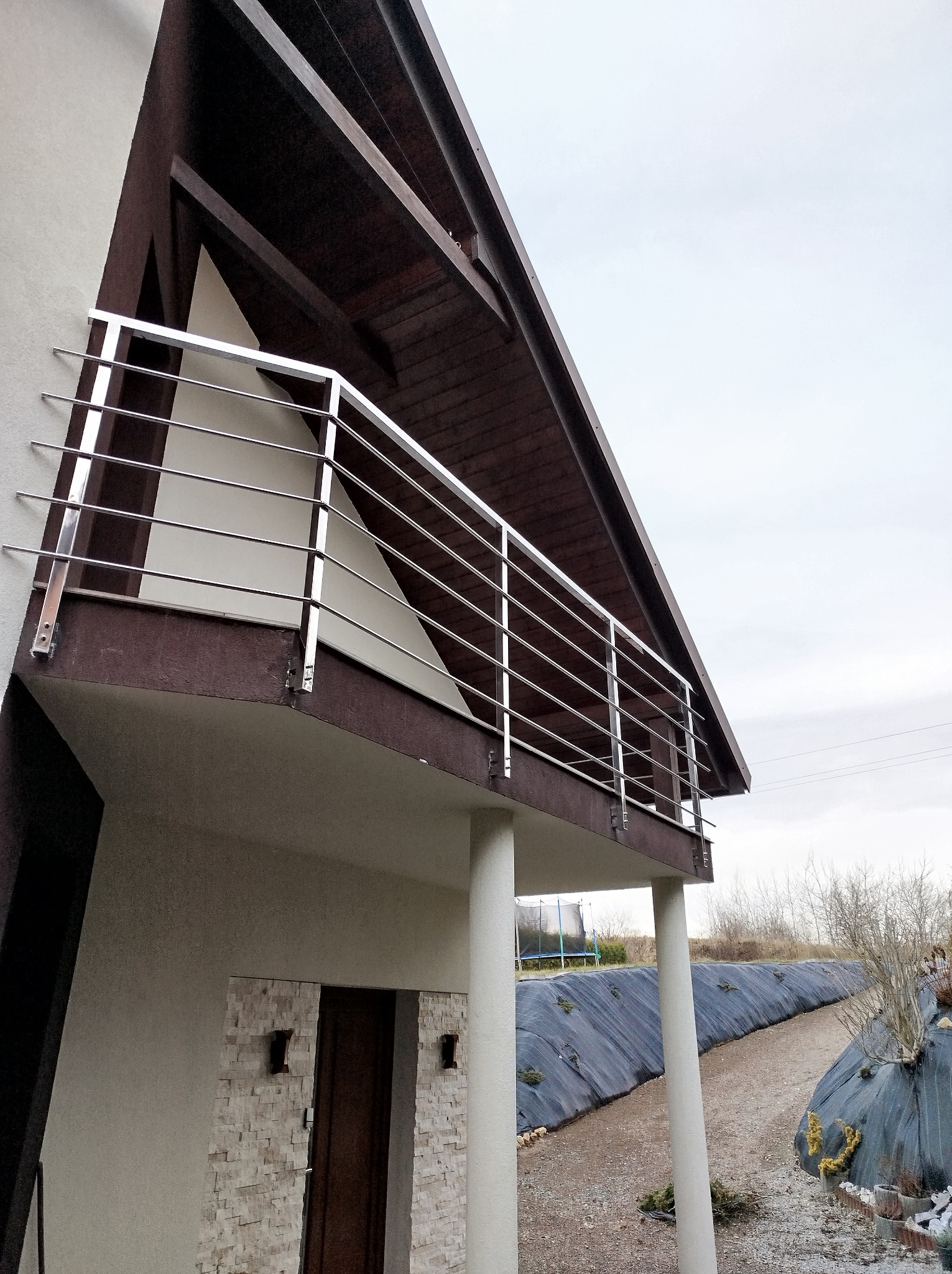 Balkon z prostą, poziomą balustradą ze stali nierdzewnej w Wadowicach, widok z dołu, na tle szarego nieba i zagospodarowanego terenu.