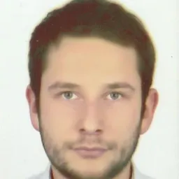 Portret młodego mężczyzny z krótkimi, ciemnymi włosami i lekkim zarostem, na jasnym tle. Ujęcie od przodu, skupione na twarzy.