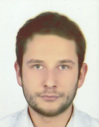 Portret młodego mężczyzny z krótkimi, ciemnymi włosami i lekkim zarostem, na jasnym tle. Ujęcie od przodu, skupione na twarzy.