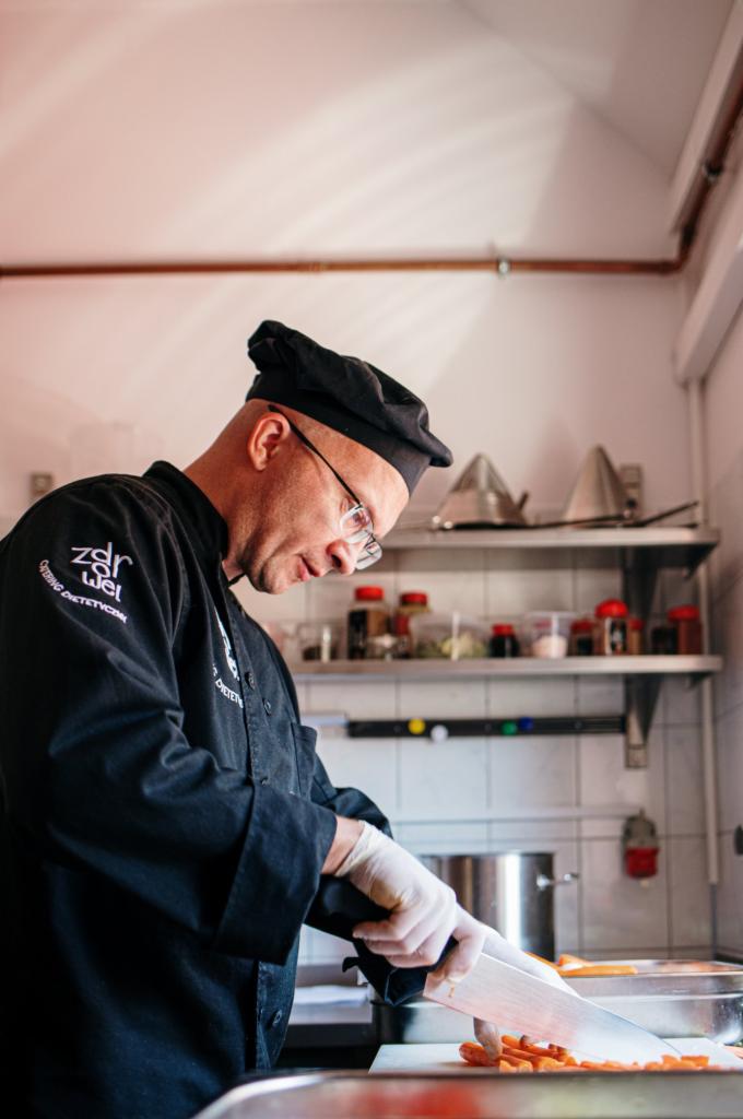 Kucharz w czarnym uniformie kroi marchewkę na desce. W tle półki z przyprawami i naczyniami. Przygotowanie posiłku zgodnie z zaleceniami.