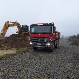Ciężarówka Mercedes-Benz Actros z włączonymi światłami mijania, na drodze z utwardzonego kruszywa, w tle żółta koparka Caterpillar na hałdzie ziemi, pochmurny dzień.