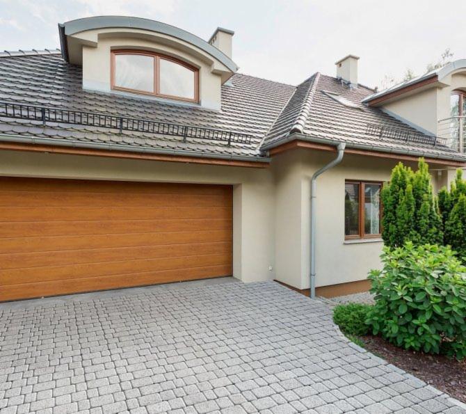 Podjazd do domu z ułożoną szarą kostką brukową w jodełkę, widoczny garaż z drewnianą bramą, fragment elewacji i zagospodarowana zieleń.