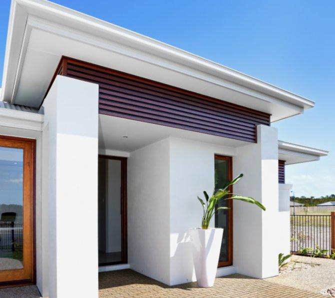 Nowoczesny dom z białymi ścianami, drewnianymi elementami dekoracyjnymi i jasną nawierzchnią przed wejściem, uzupełniony wysoką białą donicą z zieloną rośliną.