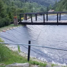 Wyłożony czarną folią wykop pod zbiornik wodny z drewnianym pomostem w ogrodzie na tle zalesionych wzgórz.