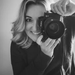 Uśmiechnięta blondynka spogląda przez obiektyw aparatu Sony Alpha 7III z dużym obiektywem, czarno-białe zdjęcie.