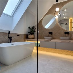 Studio Metr Na 2 - Projektowanie Wnętrz i Mebli - Stylowa łazienka na poddaszu z owalnym lustrem, drewnianą mozaiką i wolnostojącą wanną. Duże okno dachowe doświetla wnętrze, a podświetlenie szafki dodaje ciepła.