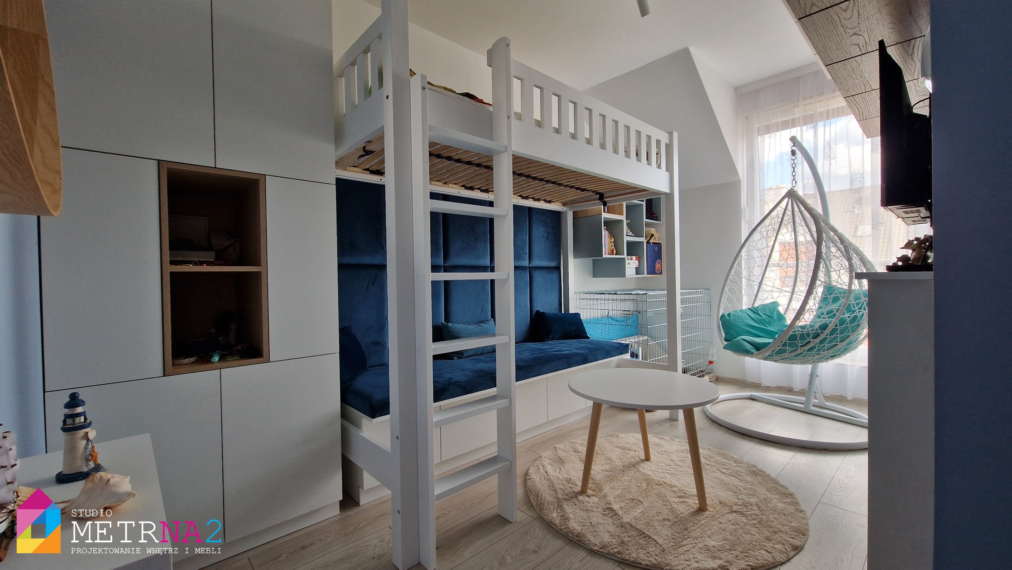 Pokój dziecięcy z łóżkiem piętrowym, zabudową meblową i wiszącym fotelem. Całość w jasnych barwach z akcentami granatu i turkusu. Widok na okno z delikatną firanką.