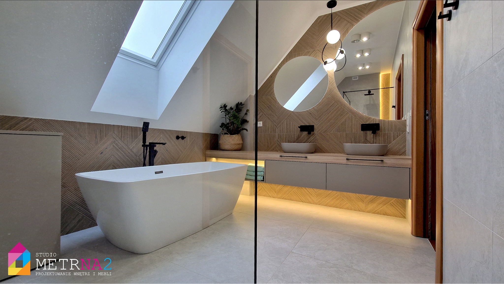 Stylowa łazienka na poddaszu z owalnym lustrem, drewnianą mozaiką i wolnostojącą wanną. Duże okno dachowe doświetla wnętrze, a podświetlenie szafki dodaje ciepła.