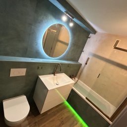Damian Bałuk - Nowoczesna łazienka z okrągłym lustrem podświetlanym LED, białą umywalką na szafce i zielonym podświetleniem podłogi. Ciemne płytki na ścianach.