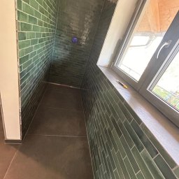 Damian Bałuk - Kabina prysznicowa z zielonymi, błyszczącymi płytkami ułożonymi w cegiełkę, brązowa podłoga i okno dachowe z widokiem na zewnątrz.