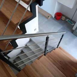 Damian Bałuk - Drewniane schody z metalową balustradą, widok z góry. Szare płytki na podłodze, czerwone wiadro i mop w tle. Minimalistyczny design, nowoczesne wykończenie.