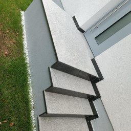 Damian Bałuk - Zewnętrzne schody z szarego kamienia z czarnymi krawędziami, prowadzące do budynku z oknem. Obok trawnik z białym obramowaniem. Ujęcie z góry, podkreślające geometrię.