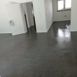 Damian Bałuk - Wykończone wnętrze z szarą, matową podłogą z płytek, białe ściany i otwarte drzwi. Widoczne gniazdka elektryczne i okno z parapetem.