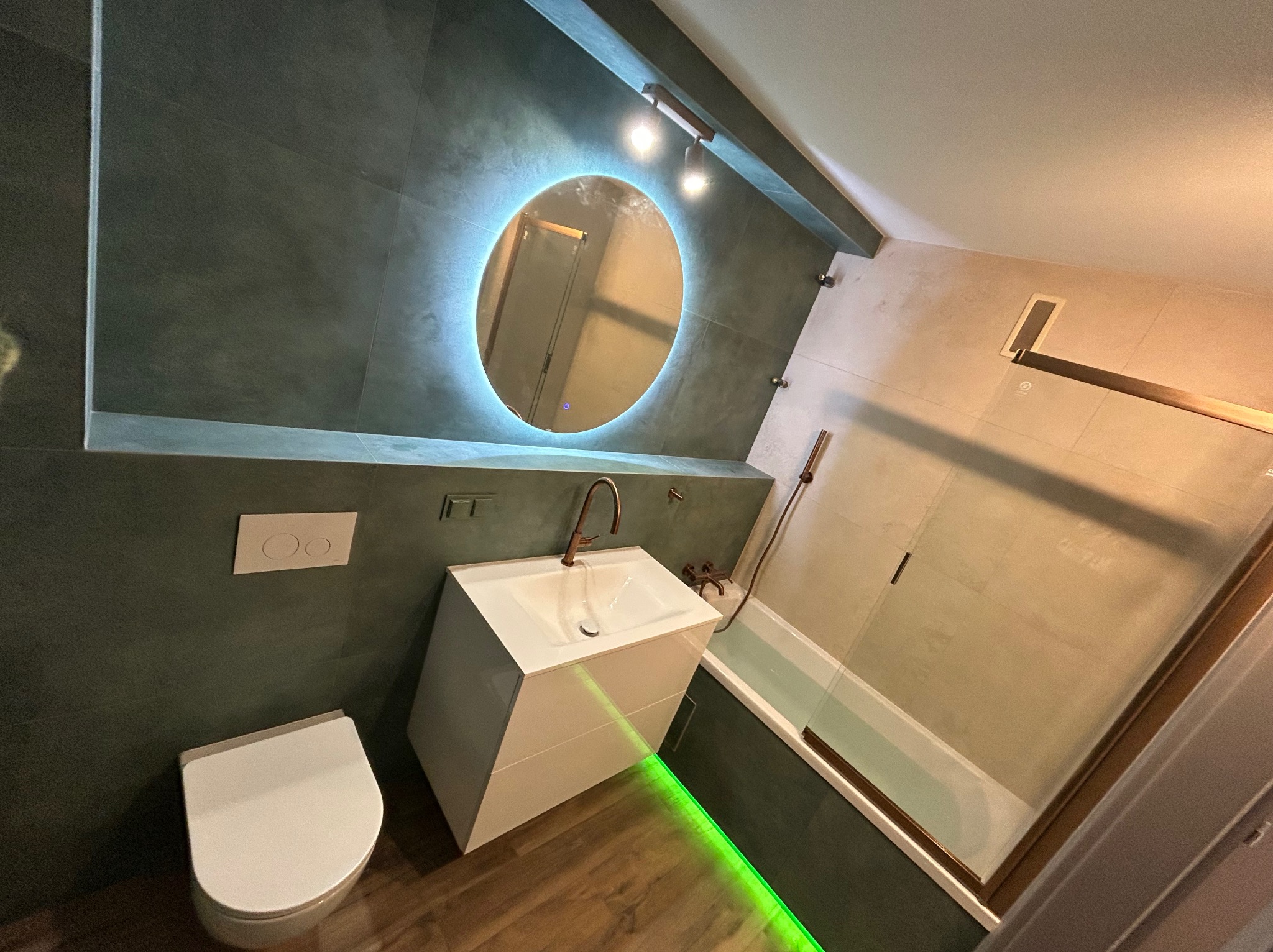Nowoczesna łazienka z okrągłym lustrem podświetlanym LED, białą umywalką na szafce i zielonym podświetleniem podłogi. Ciemne płytki na ścianach.
