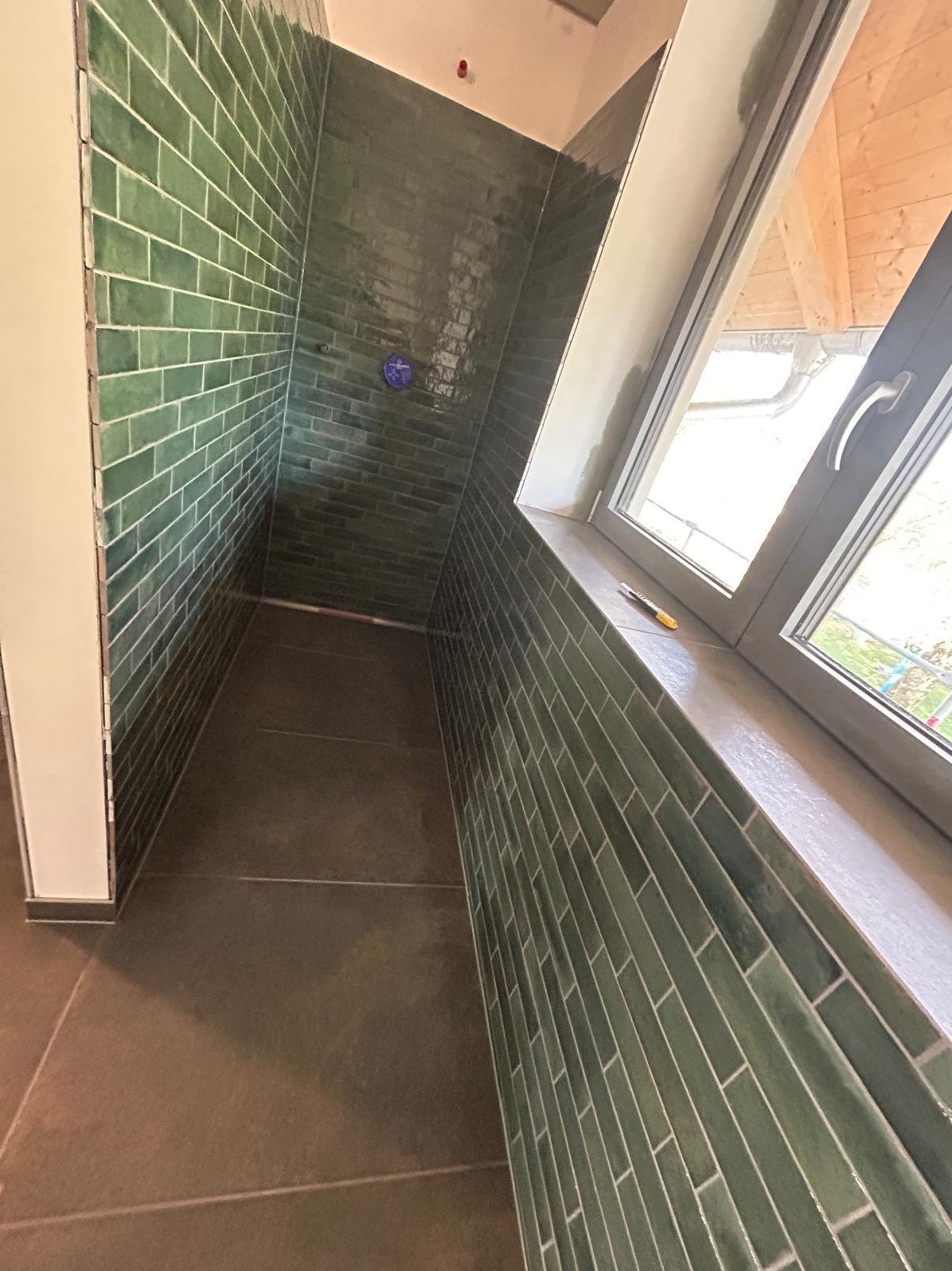 Kabina prysznicowa z zielonymi, błyszczącymi płytkami ułożonymi w cegiełkę, brązowa podłoga i okno dachowe z widokiem na zewnątrz.