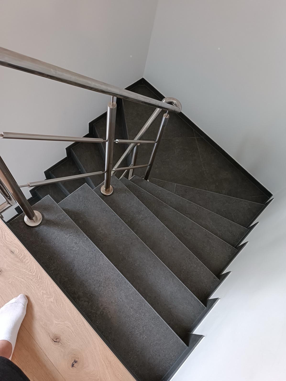 Schody wykończone ciemnoszarą płytką, z metalową balustradą, widziane z góry. Fragment drewnianej podłogi i stopy w białej skarpetce w lewym dolnym rogu.