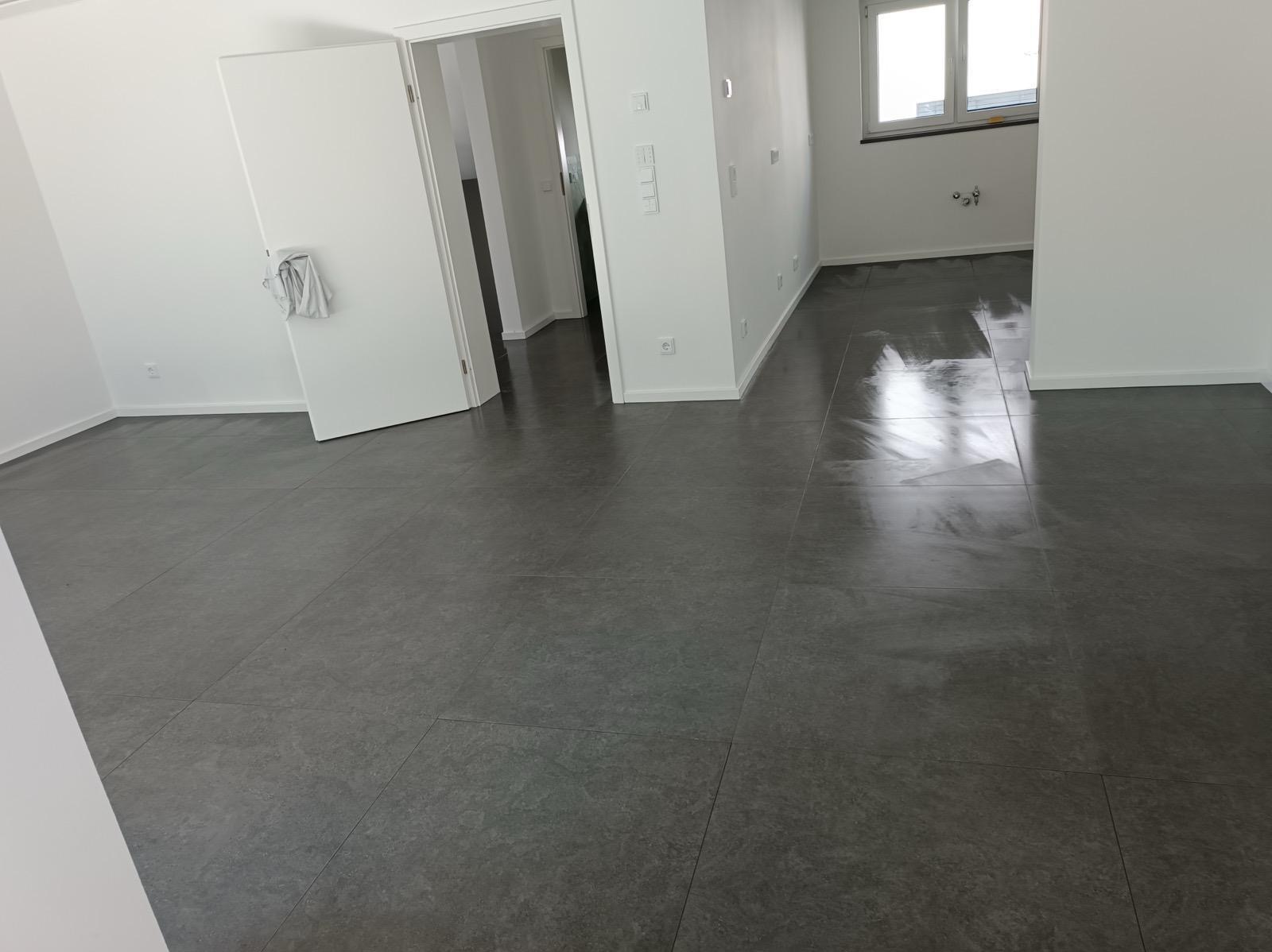 Wykończone wnętrze: szare płytki podłogowe, białe ściany, otwarte drzwi. Widoczne gniazdka elektryczne i okno. Minimalistyczny styl, świeżo po remoncie.