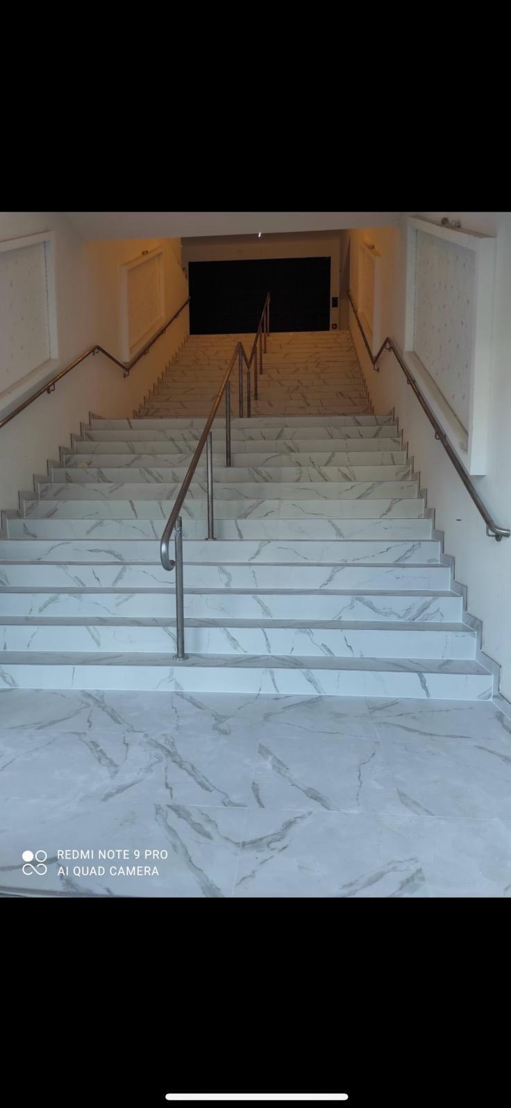 Białe marmurowe schody z metalową balustradą prowadzące w górę, oświetlone światłem z góry. Widok z dołu, symetryczny kadr.