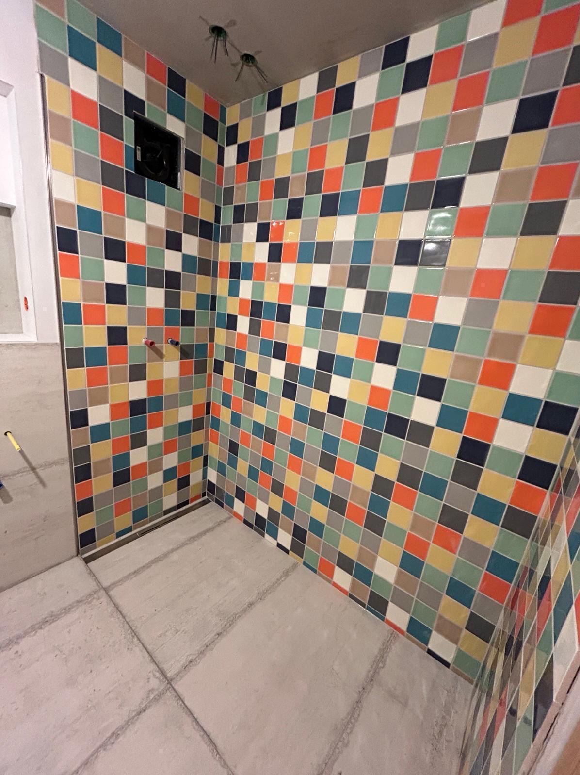 Ściana kabiny prysznicowej wyłożona kolorową, kwadratową mozaiką. Widoczny odpływ liniowy i surowy beton na podłodze. W tle fragment ściany z instalacją.
