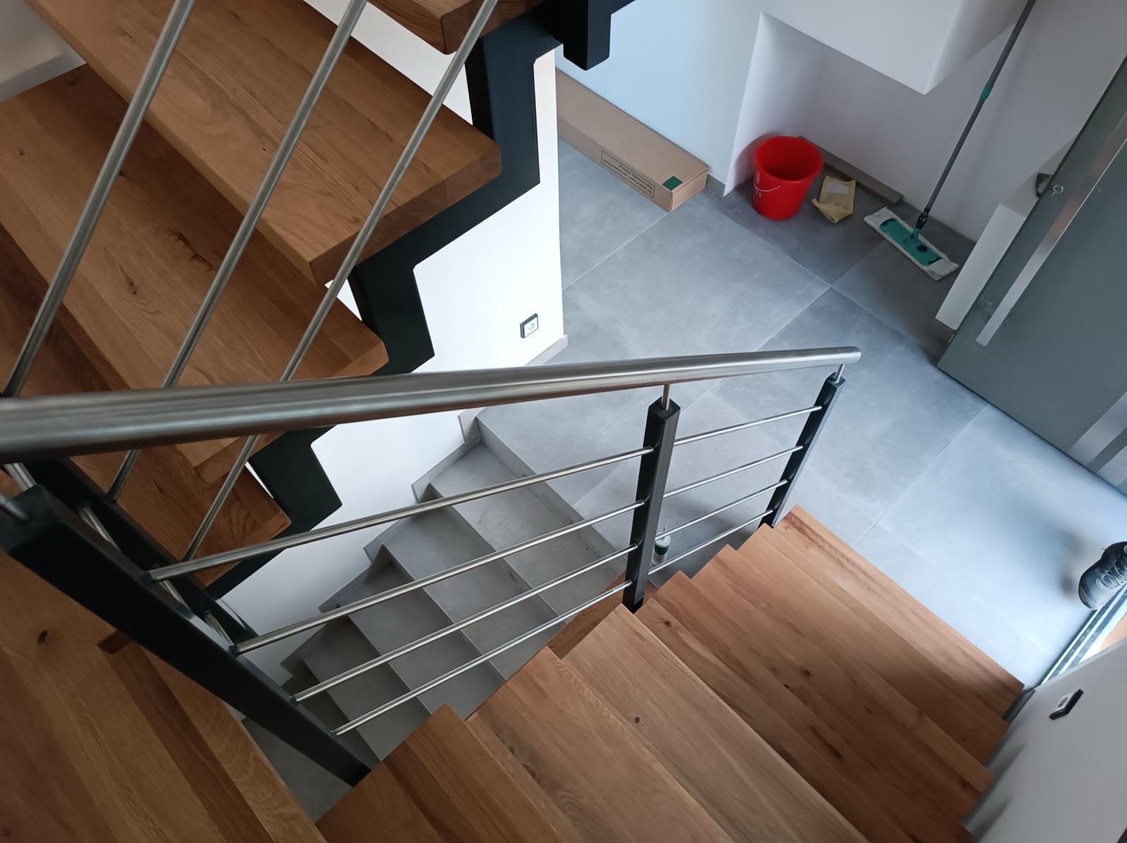 Nowoczesne schody z drewnianymi stopniami i metalową balustradą, widok z góry. Szare płytki na podłodze, czerwone wiadro i mop w tle.