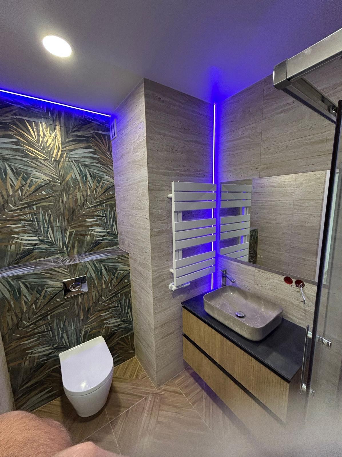 Nowoczesna łazienka z podświetleniem LED, płytkami w liście i kamienną umywalką. Grzejnik drabinkowy i toaleta wkomponowane w aranżację. Ukończony projekt.