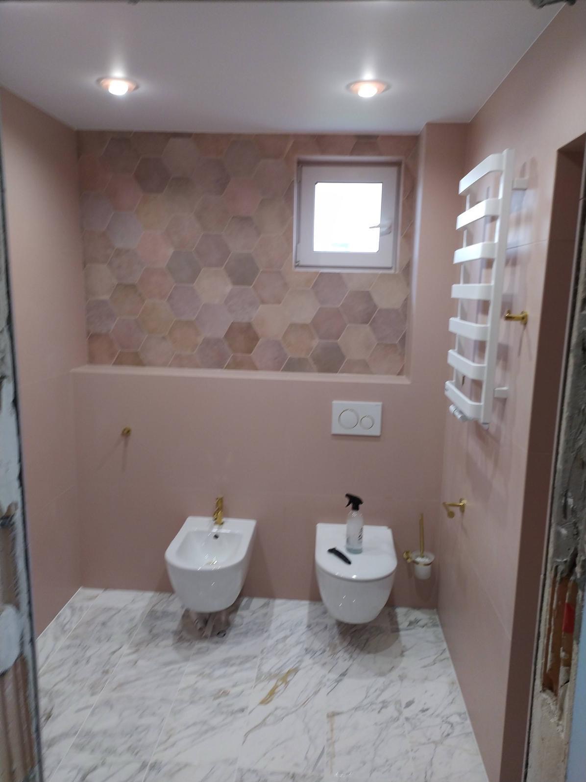 Wykończona łazienka w odcieniach różu i beżu z heksagonalnymi płytkami na ścianie, marmurową podłogą i białymi sanitariatami. Widoczny bidet, toaleta i grzejnik drabinkowy.