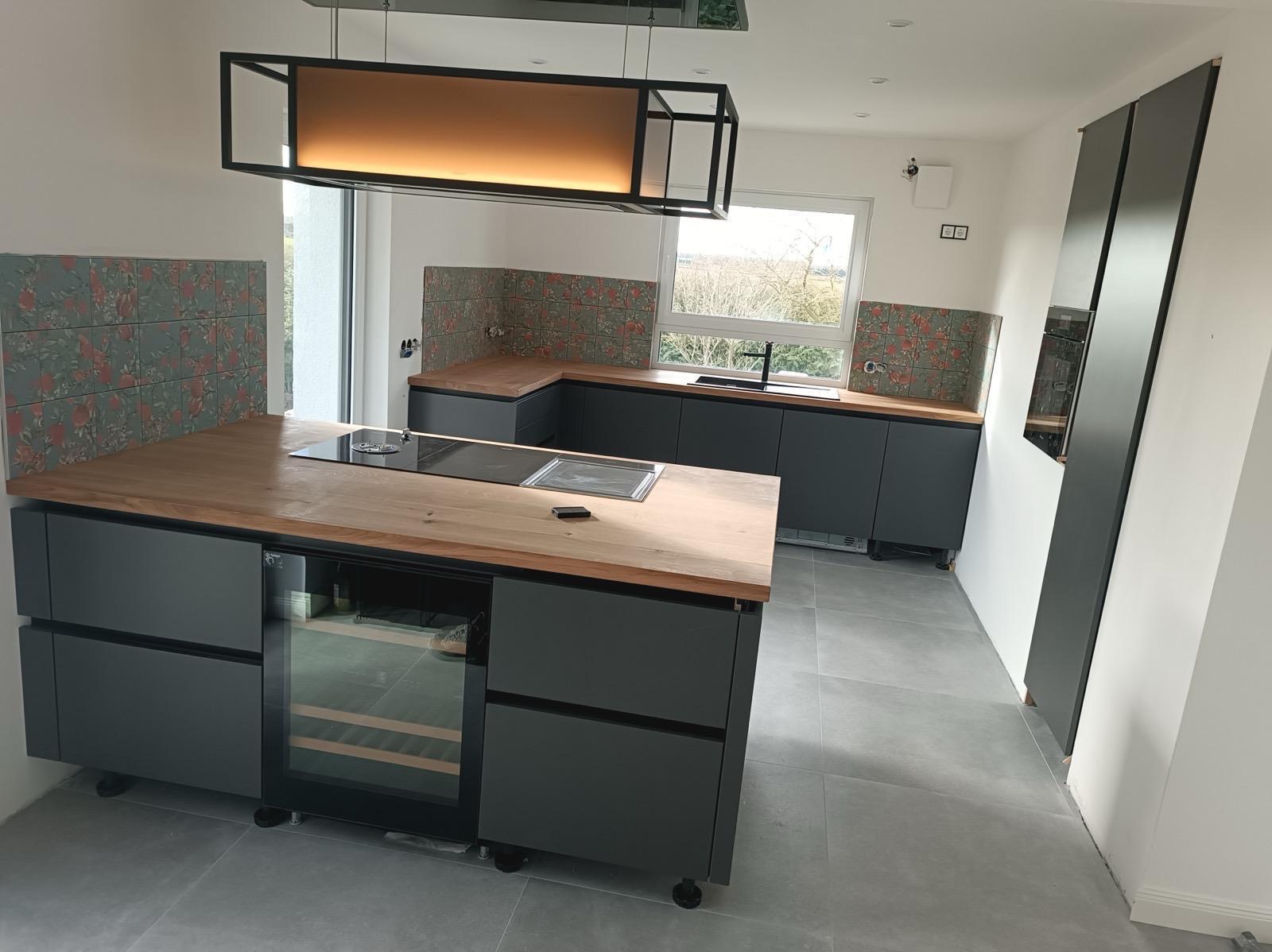 Nowoczesna kuchnia z szarymi szafkami, drewnianym blatem i wyspą z chłodziarką na wino. Nad wyspą designerska lampa. Na ścianie płytki z motywem kwiatowym.