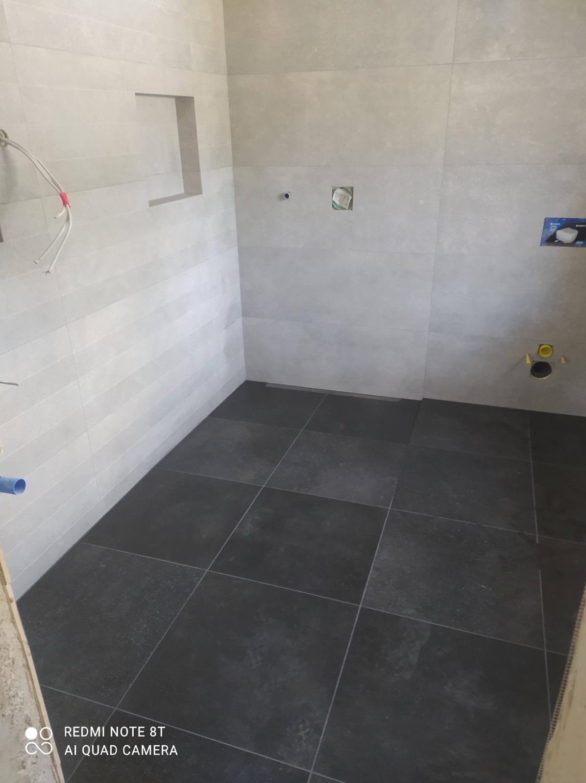 Wykończona łazienka z ciemną podłogą z płytek i jasnymi, poziomymi płytkami na ścianach. Widoczne instalacje i wnęka w ścianie. Minimalistyczny design.