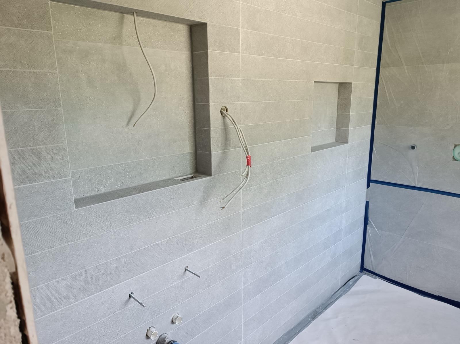 Łazienka w trakcie wykańczania: szare, poziome płytki z wnękami, wystające kable i zabezpieczona folią podłoga. Widoczne elementy instalacji wodnej.