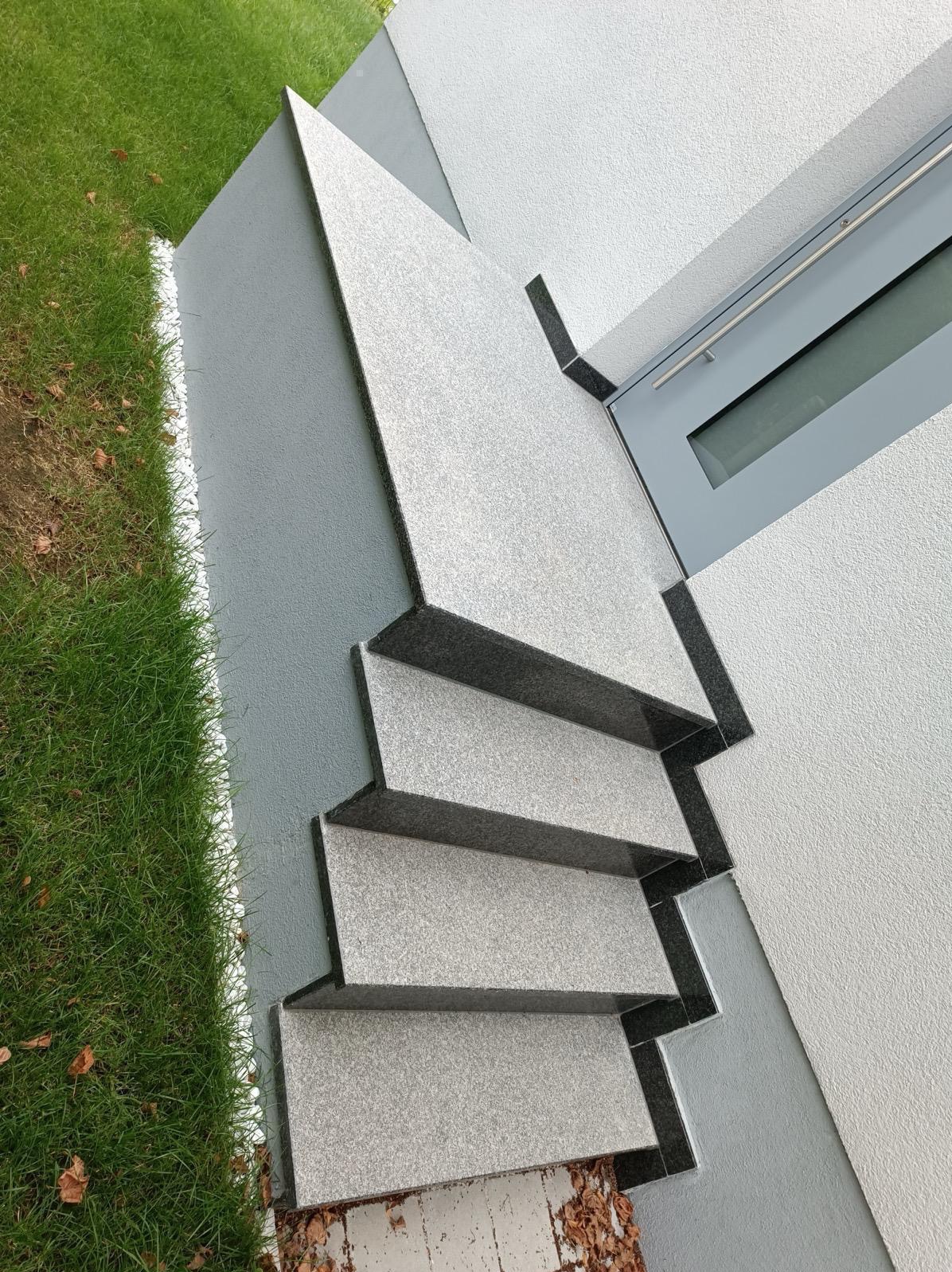 Zewnętrzne schody z szarego kamienia z czarnymi krawędziami, prowadzące do budynku z oknem. Obok trawnik z białym obramowaniem. Ujęcie z góry, podkreślające geometrię.