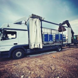 Transport Kamil Paluch - Biała ciężarówka z naczepą kurtynową, na której załadowano niebieskie agregaty prądotwórcze, rozładunek za pomocą małej koparki na gąsienicach w terenie budowy.