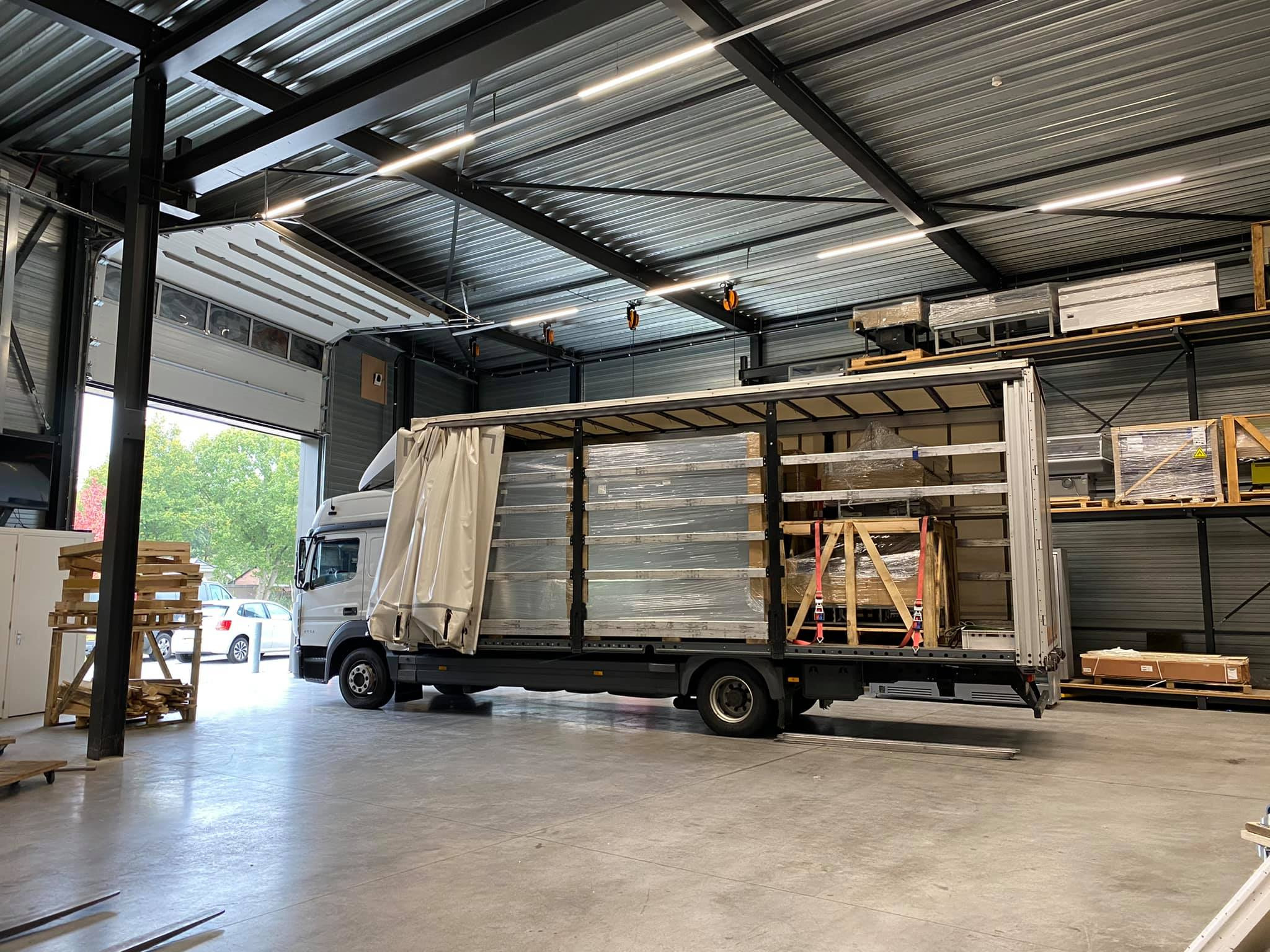 Ciężarówka z odkrytą naczepą, załadowana paletami z towarem w folii, stojąca w magazynie z regałami wypełnionymi pudełkami, widok z boku, z zewnątrz widoczny fragment parkingu.