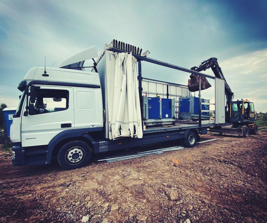 Biała ciężarówka z naczepą kurtynową, na której załadowano niebieskie agregaty prądotwórcze, rozładunek za pomocą małej koparki na gąsienicach w terenie budowy.