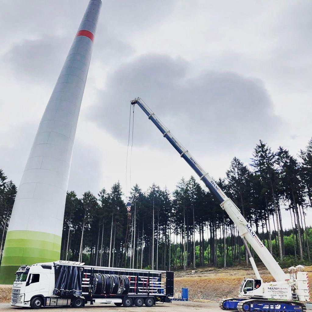 Transport turbiny wiatrowej na naczepie niskopodwoziowej, z użyciem dźwigu gąsienicowego na tle lasu iglastego i pochmurnego nieba.