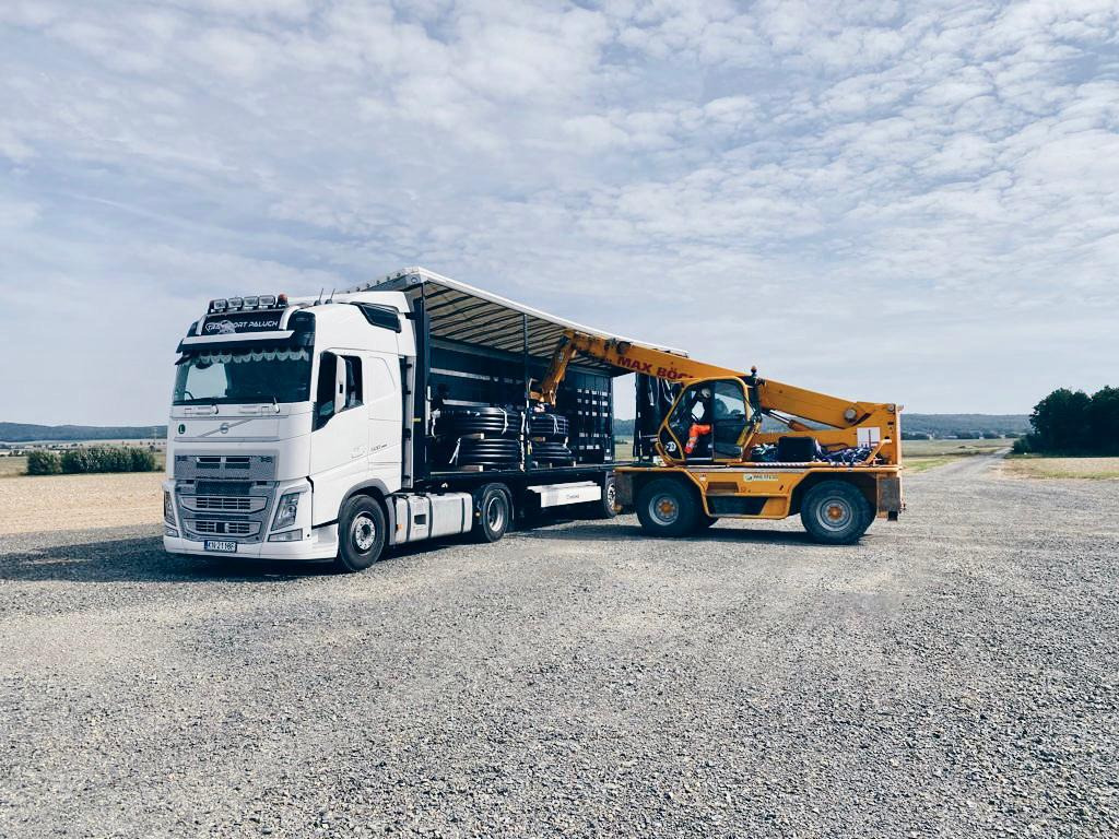 Biała ciężarówka z naczepą załadowaną czarnymi rurami, obok żółty wózek widłowy z operatorem w pomarańczowym kombinezonie, na żwirowym placu, w tle pole i las.