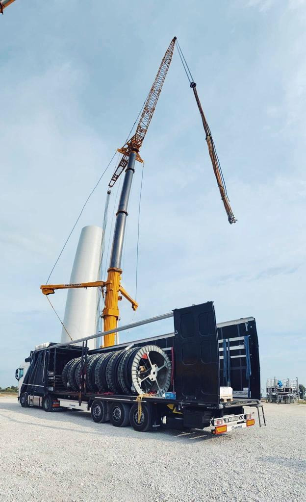 Ciężarówka z naczepą przewozi elementy turbiny wiatrowej, w tle widać dźwig budowlany i fragmenty innych turbin.