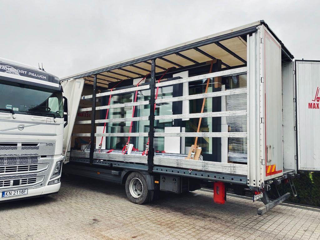 Ciężarówka z naczepą załadowana oknami, zabezpieczonymi folią i pasami transportowymi, widoczny numer rejestracyjny KN 2118F i logo firmy transportowej na kabinie.