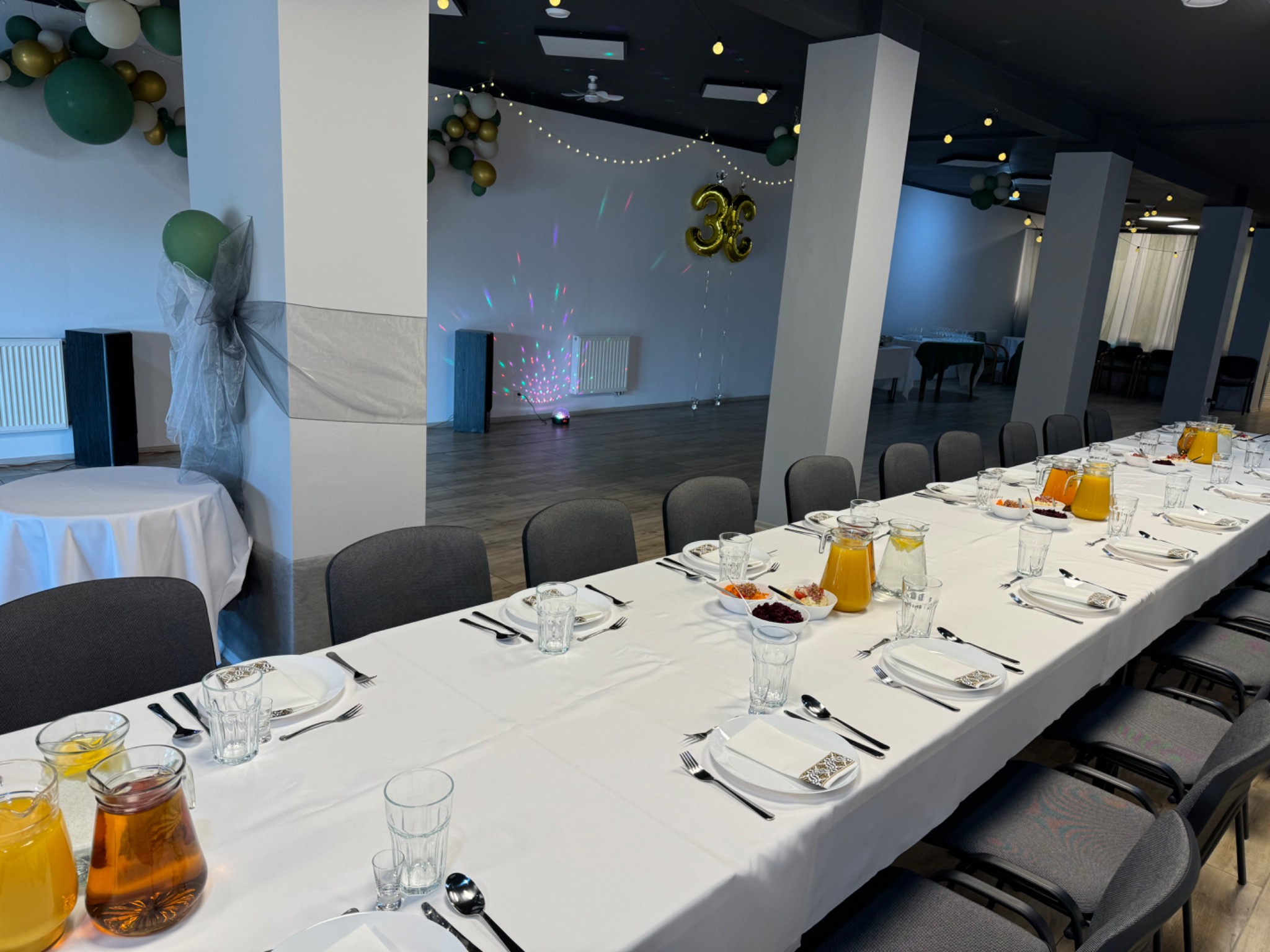 Uroczystość w przestronnej sali: długi stół z białym obrusem, nakryty zastawą i dzbankami z sokami, dekoracje balonowe w odcieniach zieleni i złota, oświetlenie punktowe i dyskotekowe.