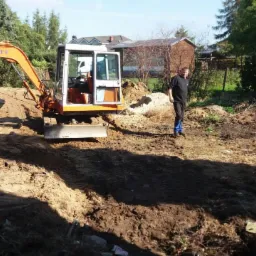 Pomarańczowa mini koparka Schaeff na placu budowy, obok stoi mężczyzna w czarnej bluzie i niebieskich spodniach, w tle zabudowania gospodarcze i drzewa.