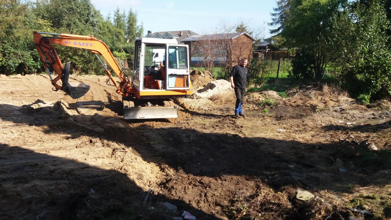 Pomarańczowa mini koparka Schaeff na placu budowy, obok stoi mężczyzna w czarnej bluzie i niebieskich spodniach, w tle zabudowania gospodarcze i drzewa.