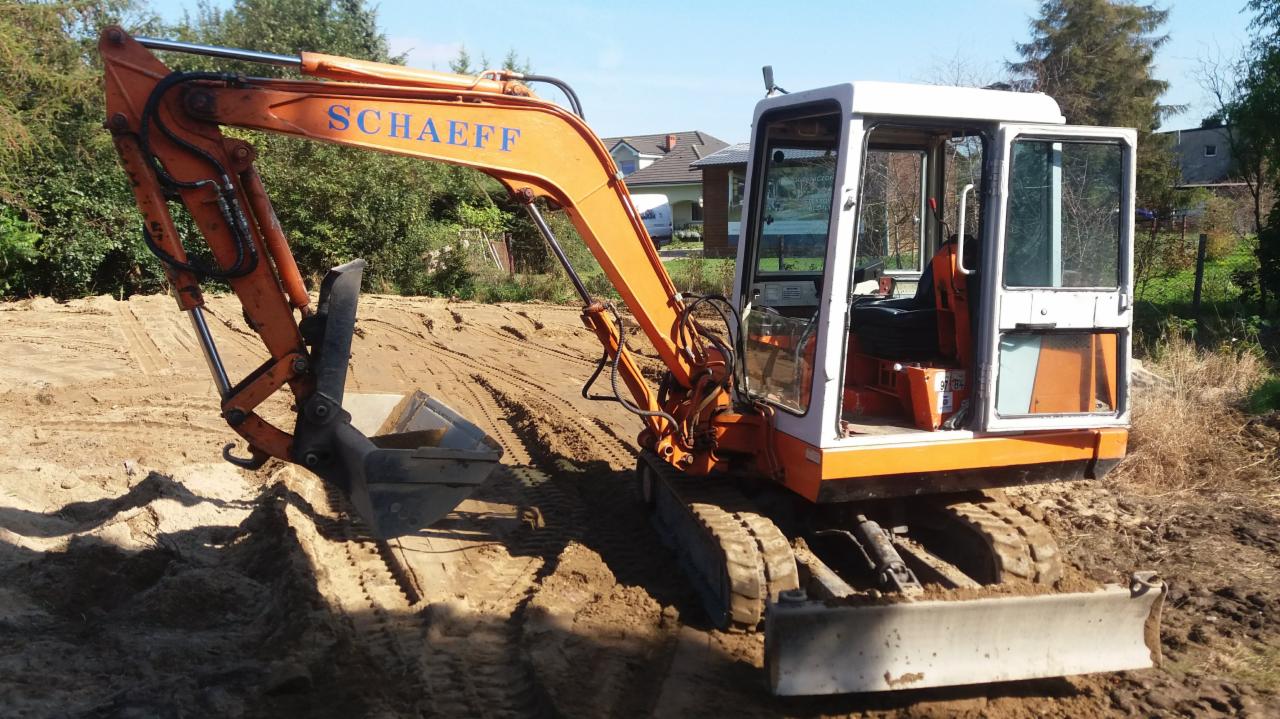 Pomarańczowo-biała minikoparka Schaeff na nierównym, piaszczystym terenie, widoczne ślady gąsienic i wykopany dół, w tle domy jednorodzinne i drzewa pod błękitnym niebem.
