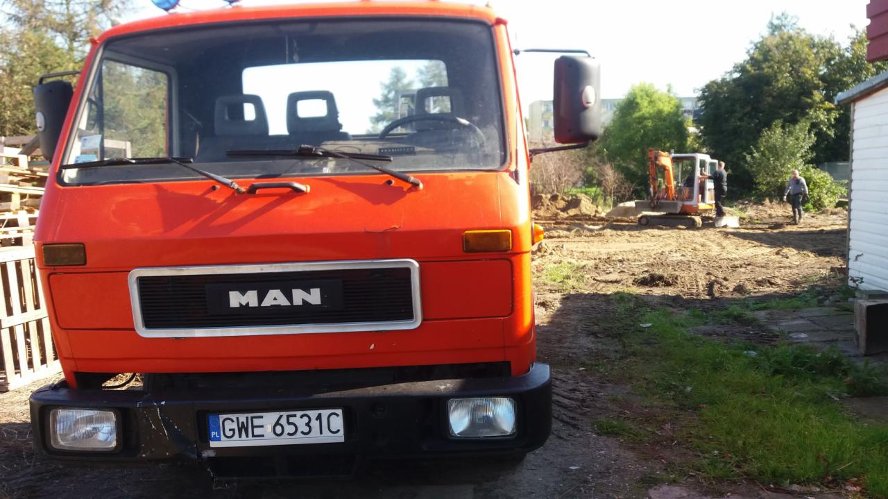 Pomarańczowa ciężarówka MAN na placu budowy z wykopem i małą koparką w tle.