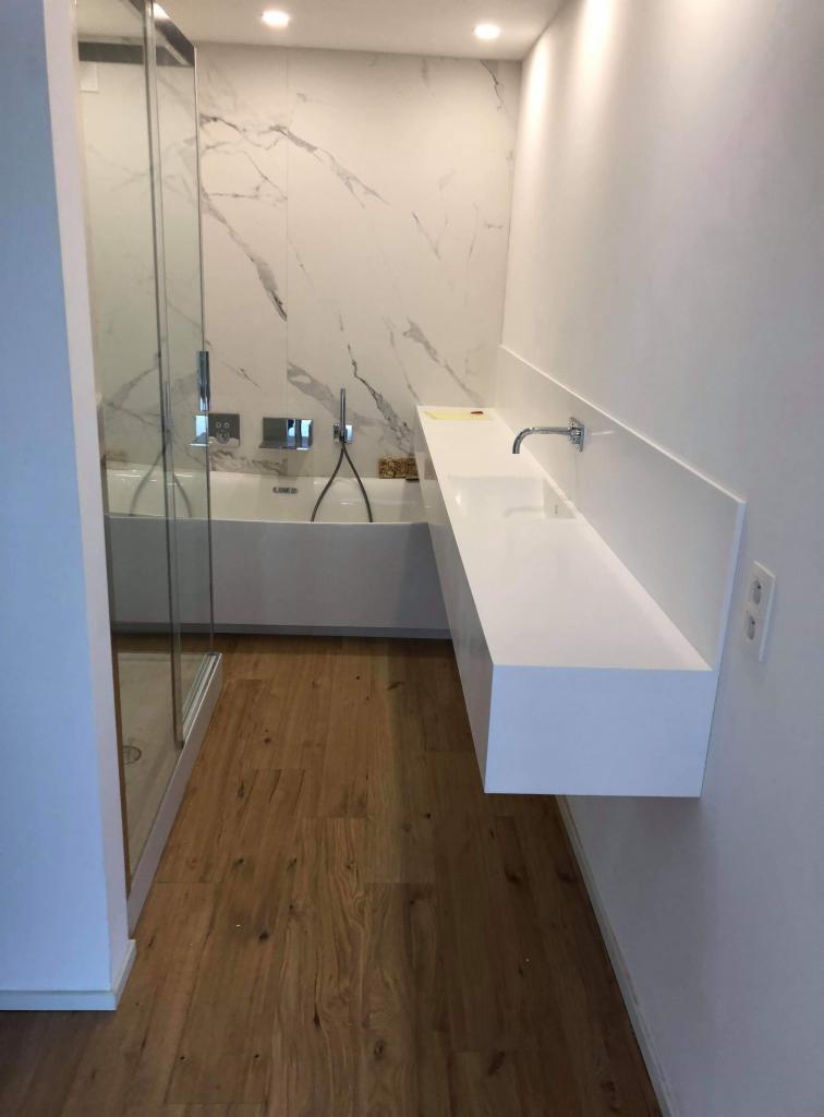 Nowoczesna łazienka z marmurową ścianą, minimalistyczną białą umywalką zawieszoną na ścianie i drewnianą podłogą, widok z perspektywy wejścia.