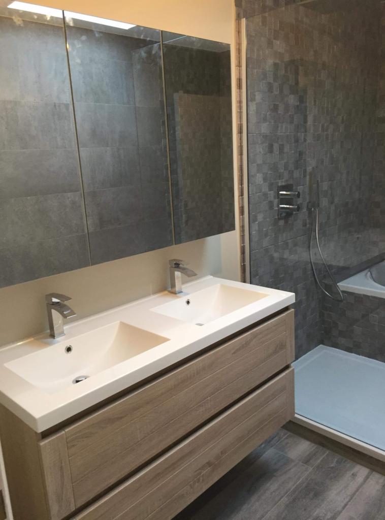 Wykończona łazienka z dwoma umywalkami nablatowymi, drewnopodobną szafką z szufladami, dużym lustrem z oświetleniem LED i kabiną prysznicową wyłożoną mozaiką w odcieniach szarości.